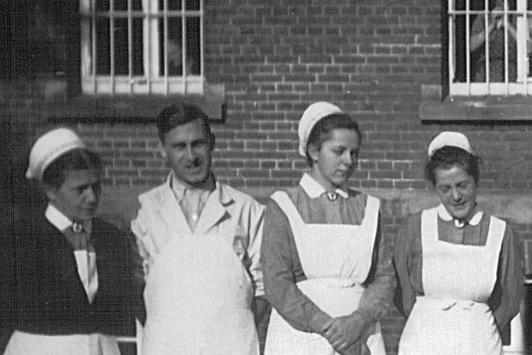 Krankenschwestern und ein Pfleger der Heil- und Pflegeanstalt Wehnen vor dem Hauptgebäude der Klinik um 1943. Im Hintergrund Patienten hinter vergitterten Fenstern.