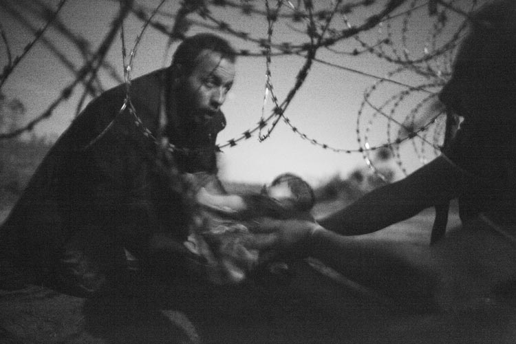 Durch ein Loch in einem Stacheldrahtzaun wird ein Baby an einen syrischen Flüchtling übergeben, der die Grenze zwischen Serbien und Ungarn in der Nähe von Röszke überwunden hat. Ungarn verhärtete zu dieser Zeit seine Haltung gegenüber Flüchtlingen, die ins Land wollten. Im Juli 2015 hatte man begonnen, einen vier Meter hohen Zaun entlang der Grenze mit Serbien zu bauen, um den Übertritt zu verhindern. Flüchtlinge versuchten, Wege zu finden, bevor der Bau des Zauns am 14. September beendet wurde. Diese Gruppe hatte sich die Nacht vier Stunden der Grenzpolizei in einem Apfelgarten versteckt. Foto: Warren Richardson, Australien, Scanpix/Panos Pictures Das Gewinnerfoto der World Press Photo Ausstellung von Warren Richardson.