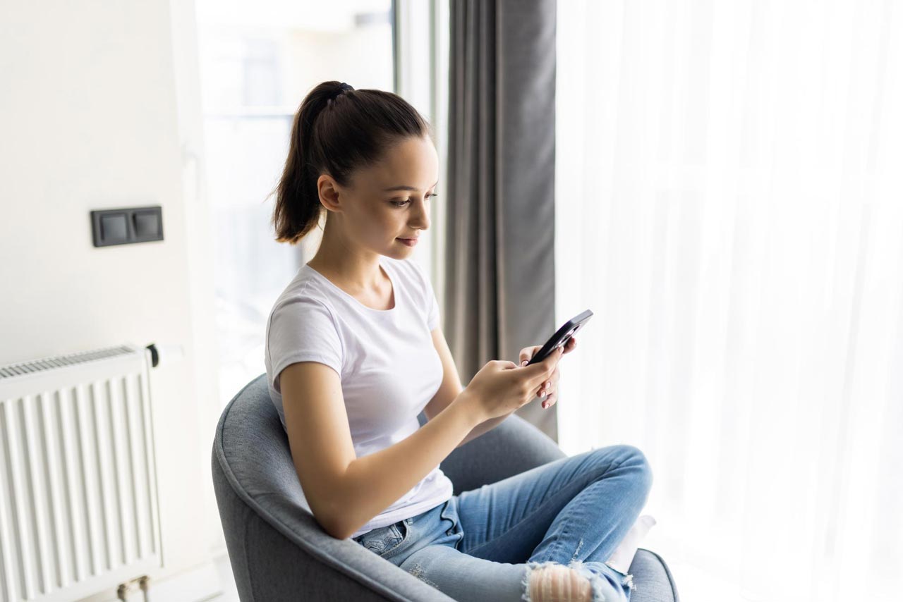 Eine junge Frau sitzt am Fenster mit Smartphone, links im Raum steht ein weißer Heizkörper.